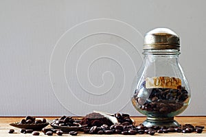 TABLE TOP IMAGE OF ROASTED COFFEE BEANS IN A CLEAR COFFEE STORAGE JAR AND WOODEN SCOOP ON WOODEN SURFACE
