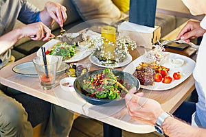 At the table, two men eat dinner, eat a steak, with a salad on a white plate, with a fork and knife in their hands.