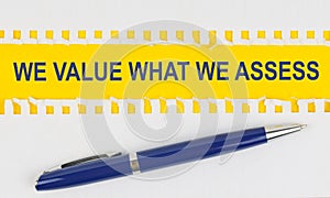 On the table are two sheets from a notebook and a pen on a yellow background written - WE VALUE WHAT WE ASSESS