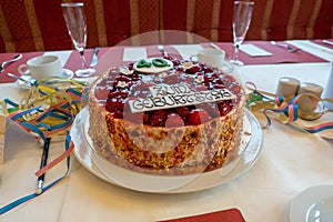Table with a white tablecloth there is a red birthday strawberry cake with the inscription