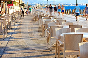 The tables and chairs of the beach cafe are located along the promenade among people walking near the blue sea in blur