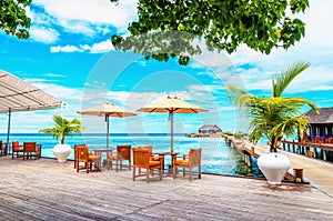 Tables and chairs with sun umbrellas on a wooden pier against the azure water of the ocean and wooden bungalows on the