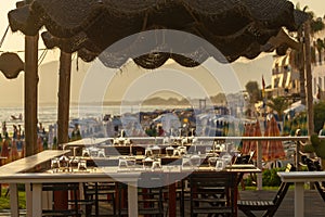 Tables in outdoor cafe or restaurant served for dinner on crowded beach with sea view, summer holidays