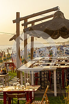 Tables in outdoor cafe or restaurant served for dinner on crowded beach with sea view, summer holidays