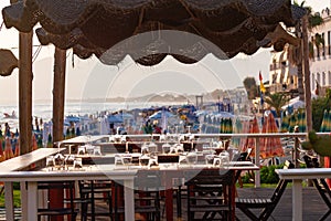 Tables in outdoor cafe or restaurant served for dinner on crowded beach with sea view, summer holidays