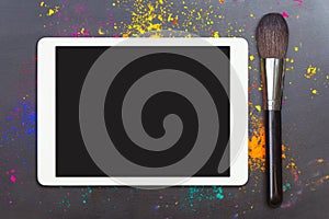 Tablet computer with blank screen with makeup brush on black background with colorful cosmetic powder. Flat lay