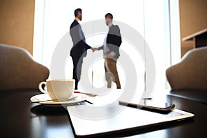 Tablet computer, coffee and smartphone with businessmen handshaking on background