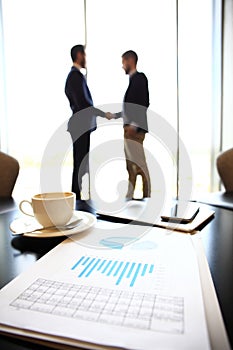 Tablet computer, coffee and smartphone with businessmen handshaking on background