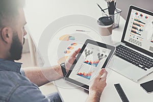 Tablet computer with graphs, diagrams and charts on screen in hands of young bearded businessman sitting at table