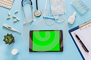 Tablet computer with green screen on blue background. Top view of doctor or nurse desk with various medical supplies and