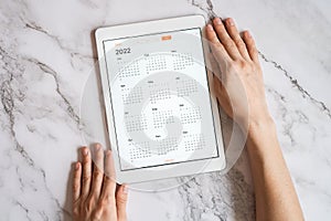 Tablet computer with an open app of calendar for 2022 year in a womans hands on a gray marble background. concept business or to d