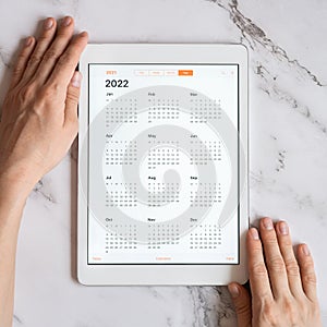 Tablet computer with an open app of calendar for 2022 year in a womans hands on a gray marble background. concept business or to d