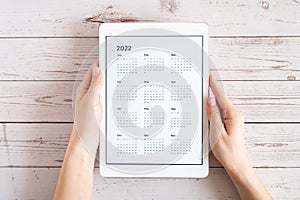 tablet computer with an open app of calendar for 2022 year in a womans hands on a wooden boards background. concept business or to