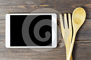 A tablet computer and wooden spoon and fork on a brown background. Copy space. Concept of cooking recipes