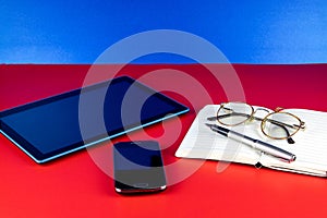 Tablet Device with Mobile Phone Notebook Pen and Spectacles on a Red Surface and Blue Background