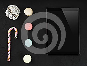 Tablet in high definition with colorful macarons, sweet lollipop and biscuit on stone black background. Copy space