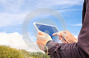 Tablet in nature outdoors. Farm or garden with blue sky and clouds. Man using smart device outside. Green grass field or park.