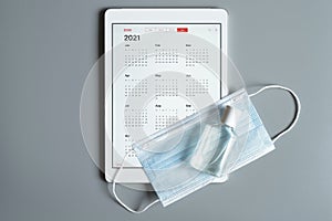 a tablet with an open calendar for 2021 year and protective medical mask and hand sanitizer on a gray background. covid-19 coronav