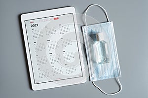 a tablet with an open calendar for 2021 year and protective medical mask and hand sanitizer on a gray background. covid-19 coronav