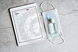 A tablet with an open calendar for 2021 year and protective medical mask and hand sanitizer on a gray concrete background. covid-1