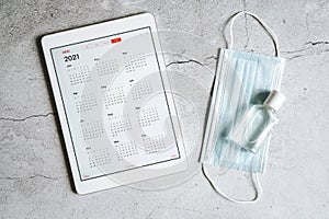 a tablet with an open calendar for 2021 year and protective medical mask and hand sanitizer on a gray concrete background. covid-1