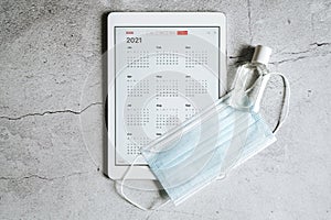 a tablet with an open calendar for 2021 year and protective medical mask and hand sanitizer on a gray concrete background. covid-1