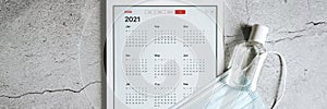 a tablet with an open calendar for 2021 year and protective medical mask and hand sanitizer on a gray concrete background. covid-1