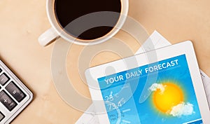 Tablet pc showing weather forecast on screen with a cup of coffee on a desk