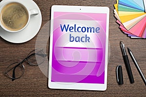 Tablet with phrase Welcome Back on wooden table. Office desk with cup of coffee, flat lay photo
