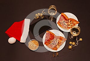 Tablet, Santa hat, slices of pepperoni pizza, ham pizza and chicken pizza, glasses of white wine, peanuts and jingle bells.
