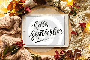 The tablet says the word hello September with red leaves and a dais on the wooden background. Concept of the autumn.