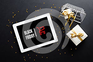 Tablet with sign `Black Friday` on screen, shopping basket, gift box, confetti over black background. Flat lay, top view, overhe