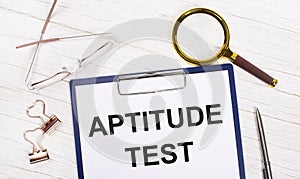 A tablet with the text APTITUDE TEST on white paper, a magnifier, glasses, golden paper clips and a pen lie on a white office