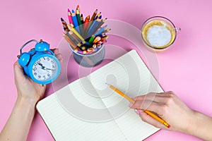 Tablet with white Leaf, next to which is a green plant, a glass with pencils. Hand holds a pen and writes a clock on the sheet in