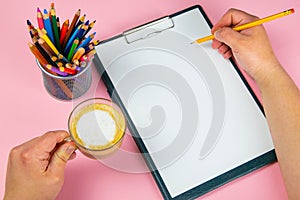 Tablet with white Leaf, next to which is a green plant, a glass with pencils. Hand holds a pen and writes a cup of coffee on the