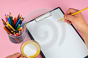 Tablet with white Leaf, next to which is a green plant, a glass with pencils. Hand holds a pen and writes a cup of coffee on the