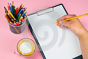 Tablet with white Leaf, next to which is a green plant, a glass with pencils. Hand holds a pen and writes a cup of coffee on the