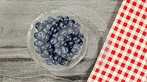 Tabletop view, small glass bowl of blueberries, red checkered gingham tablecloth next to it on gray wood desk
