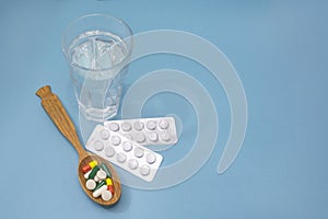 Tablets and vitamins in spoon on blue background. Transparent glass of water for medication. The concept of pharmaceuticals,