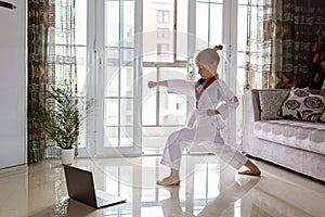 Taekwondo girl in kimono with white belt exercising at home in living room. Online education during coronavirus covid-19 lockdown