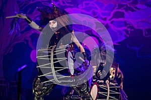 Taiko drummer in a wig and a demon mask performs on stage with drum on a dark background.