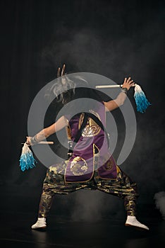 Taiko drummer in a wig and a demon mask on stage with drumsticks. Back amd profile full length portrait.