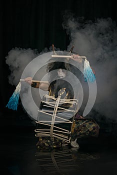 Taiko drummer in a wig and a demon mask on stage sitting with drumstick and drum