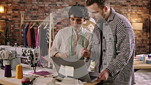 A tailor man is checking up with his seamstress on some work he s doing on the laptop, she looks very eager to help him