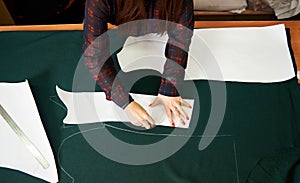 Tailor worktable in studio. Female hands making chalk markings of paper patterns on green fabric.