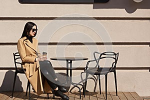 Take away coffee in women`s hands. Beautiful young brunette woman wearing brown coat sitting at table in street outdoor cafe and