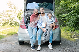 Take break at journey. Happy glad millennial islamic male and female in hijab holding baby, sitting in car trunk