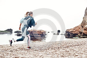 Take care of your body. Side view of strong disabled woman in sportswear with prosthetic leg is running on the beach.