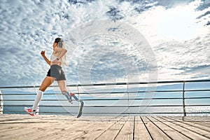 Take care of your body. Side view of a strong disabled woman in sportswear with a prosthetic leg is running on the bridge