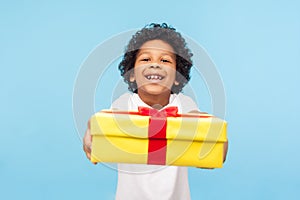 Take it! Full of joy amazing cute little boy with curly hair giving gift box to camera with excited smile